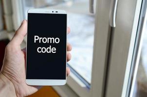 A person sees a white inscription on a black smartphone display that holds in his hand. Promo code photo