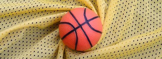 Small orange rubber basketball lies on a yellow sport jersey clothing fabric texture and background with many folds photo