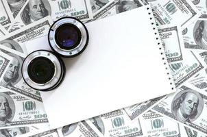 Two photographic lenses and white notebook lie on the background of a lot of dollar bills. Space for text photo