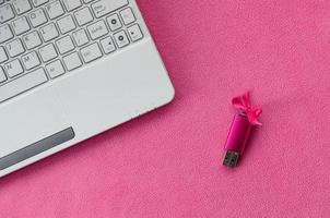 Brilliant pink usb flash memory card with a pink bow lies on a blanket of soft and furry light pink fleece fabric beside to a white laptop. Classic female gift design for a memory card photo