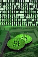 Two bitcoins lies on a pile of dollar bills on the background of a monitor depicting a binary code of bright green zeros and one units on a black background. Low key lighting photo