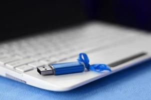 Brilliant blue usb flash memory card with a blue bow lies on a blanket of soft and furry light blue fleece fabric beside to a white laptop. Classic female gift design for a memory card photo