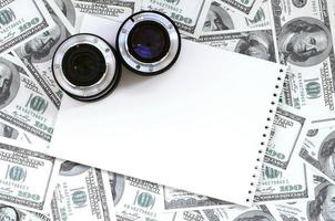 dos lentes fotográficos y un cuaderno blanco se encuentran en el fondo de muchos billetes de dólar. espacio para texto foto