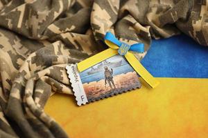 TERNOPIL, UKRAINE - SEPTEMBER 2, 2022 Famous Ukrainian postmark with russian warship and ukrainian soldier as wooden souvenir on army camouflage uniform and national flag photo