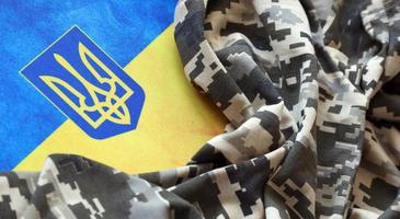 Ukrainian flag and coat of arms with fabric with texture of pixeled camouflage. Cloth with camo pattern in grey, brown and green pixel shapes with Ukrainian trident sign photo