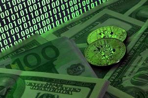 Two bitcoins lies on a pile of dollar bills on the background of a monitor depicting a binary code of bright green zeros and one units on a black background. Low key lighting photo