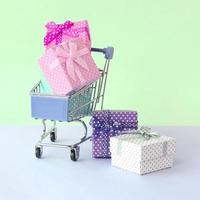 pequeñas cajas de regalo de diferentes colores con cintas en carrito de compras sobre un fondo violeta y azul pastel foto