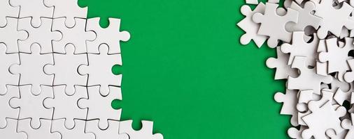 Fragment of a folded white jigsaw puzzle and a pile of uncombed puzzle elements against the background of a green surface. Texture photo with space for text