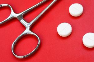 Several white tablets and a medical clamp lie on a bright red background surface. Background image on medical and pharmaceutical topics photo