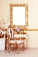 A fragment of a retro interior with a chair and table, on which is a telephone and a vase of flowers. Above the table hangs an empty picture frame for your photo