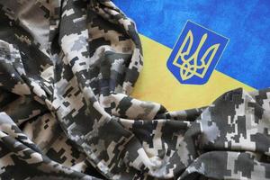 SUMY, UKRAINE - MARCH 20, 2022 Ukrainian flag and coat of arms with fabric with texture of pixeled camouflage. Cloth with camo pattern in grey, brown and green pixel shapes with Ukrainian trident sign photo