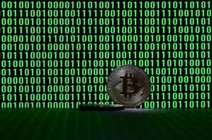 A pair of bitcoins lies on a cardboard surface on the background of a monitor depicting a binary code of bright green zeros and one units on a black background. Low key lighting photo