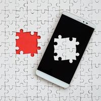 A modern big smartphone with several puzzle elements on the touch screen lies on a white jigsaw puzzle in an assembled state with missing elements photo