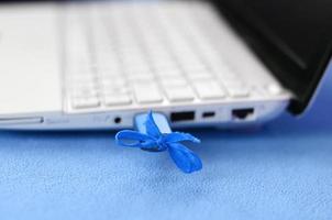 A brilliant blue USB flash drive with a blue bow is connected to a white laptop, which lies on a blanket of soft and fluffy light blue fleece fabric. Classic female design for a memory card photo