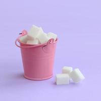 un cubo rosa en miniatura lleno de cubos de azúcar se encuentra sobre un fondo de color púrpura pastel. concepto mínimo foto
