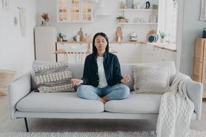 Female practice yoga, relax, sitting on sofa in lotus pose at home. Healthy lifestyle, stress relief photo