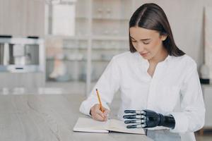 niña con discapacidad escribiendo una lista de tareas en un cuaderno planificando objetivos usando un brazo protésico biónico foto