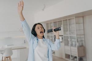 Energetic female in headphones sing karaoke, using TV remote control like a mic, rests at home photo