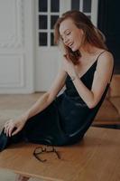 Shy beautiful lady in black silk evening dress is sitting on table at home. Concept of femininity. photo