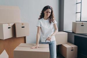 Tired hispanic woman packs parcels boxes using duct tape, frowning. Overtime, delivery service photo