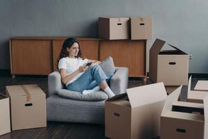 Female orders moving service using phone app on relocation day sitting in room with carton boxes photo