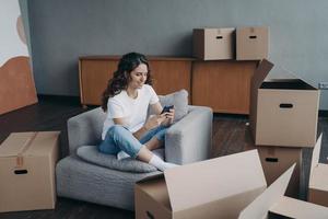 Girl uses phone app, orders moving company, shopping online for new home, sitting with carton boxes photo