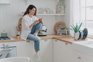 Female shopping groceries online by mobile phone app, sitting in modern kitchen at home. E commerce photo