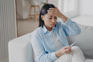 Unhealthy female suffer from fever check body temperature, holding thermometer, sitting on sofa photo