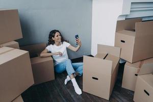 Smiling girl chatting by video call, discussing relocation details holding smartphone on moving day photo