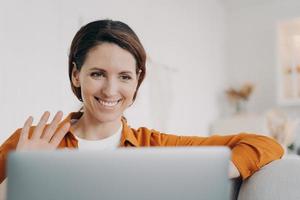 mujer hispana sonriente chateando en línea por videollamada en una laptop saludando con la mano hola, saludando a un amigo foto