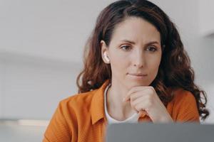 Frowning hispanic female businesswoman in earbud works at laptop, solving hard working problem photo