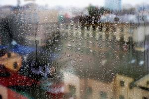 Rainy window, raindrops on the glass, against the background of a city street with houses and a road. Soft blurred background. photo