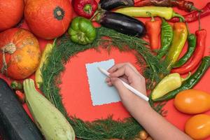 las verduras se disponen alrededor de una hoja de papel y un lápiz. espacio vacío para texto. mano femenina escribiendo una receta en un espacio en blanco vacío sobre un fondo rojo. foto