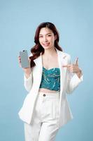 Half body of confident beautiful Asian Business woman wearing white suit smile and showing or presenting mobile phone application on hand on isolated blue background. Blank screen smartphone concept photo