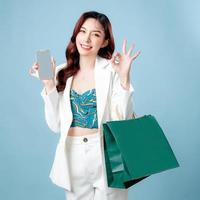 Half body of beautiful Asian Business woman wearing white suit show OK sign holding mobile phone application and shopping bag on isolated blue background. Blank screen smartphone concept. photo