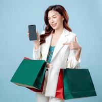 Half body of beautiful Asian Business woman wearing white suit pointing to mobile phone application and holding shopping bag on isolated blue background. Blank screen smartphone concept. photo