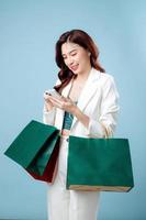 Half body of confident beautiful Asian Business woman wearing white suit using mobile phone application on hand and holding shopping bag on isolated blue background. Blank screen smartphone concept. photo