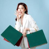 Half body of beautiful Asian Business woman wearing white suit holding shopping bag and mobile phone on isolated blue background. Blank screen smartphone concept. photo