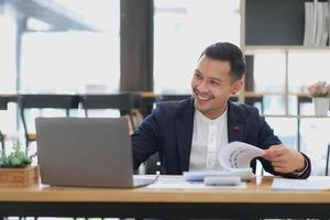 Portrait of a young Asian businessman smiling while using a laptop and writing down notes while sitting at his desk in a modern office photo