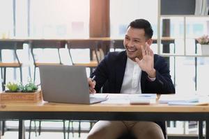Portrait of a young Asian businessman smiling while using a laptop and writing down notes while sitting at his desk in a modern office photo