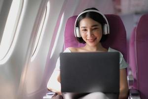 Asian young woman wearing headphone using laptop sitting near windows at first class on airplane during flight, Traveling and Business concept photo