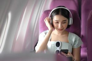 Travel tourism with modern technology and air flights concept, woman sitting in plane with modern smartphone and searching favourite music playlist in application for listening photo