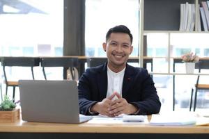 Portrait of a young Asian businessman smiling while using a laptop and writing down notes while sitting at his desk in a modern office photo