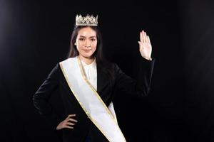 retrato de medio cuerpo del concurso de belleza miss pageant en traje inteligente asiático blazer vestido de camisa con marco de corona de diamante plateado, maquillaje de moda mano de onda de sonrisa, iluminación de estudio fondo negro aislado foto