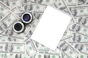 Two photographic lenses and white notebook lie on the background of a lot of dollar bills. Space for text photo