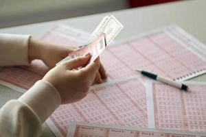 una mujer joven sostiene el billete de lotería con una fila completa de números y billetes de dólar en el fondo de las hojas en blanco de la lotería foto