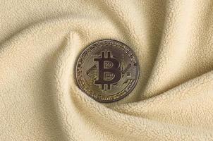 The golden bitcoin lies on a blanket made of soft and fluffy light orange fleece fabric with a large number of relief folds. The shape of the folds resembles a fan from a video card cooler photo