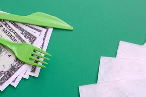 Disposable plastic cutlery green. Plastic fork and knife lie on a small amount of US dollars next to napkins photo