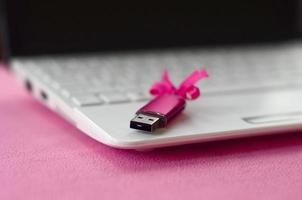 Brilliant pink usb flash memory card with a pink bow lies on a blanket of soft and furry light pink fleece fabric beside to a white laptop. Classic female gift design for a memory card photo