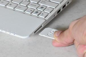 A male hand inserts a white compact SD card into the corresponding input in the side of the white netbook. Man uses modern technologies to store memory and digital data photo